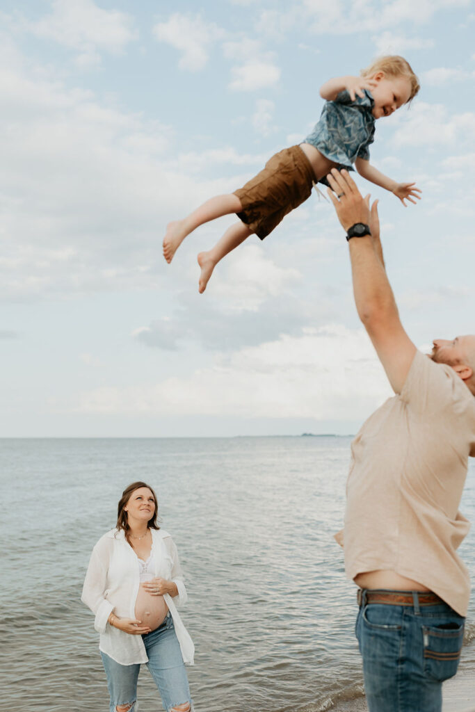 Father throwing son in the air during their Michigan beach maternity photos on Lake Huron